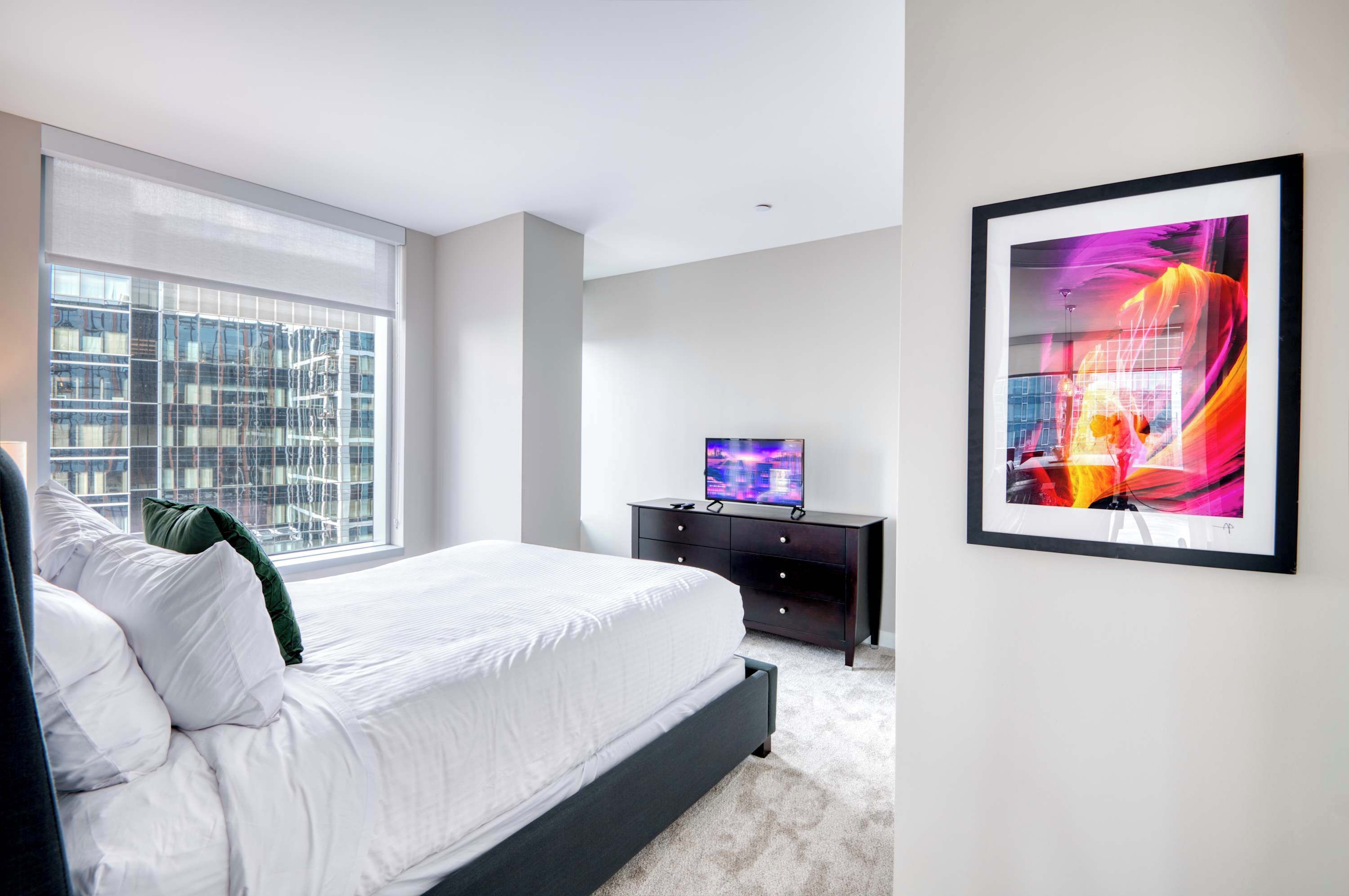 A modern bedroom with a large bed, a TV on a black dresser, and a large window with a city view.
