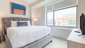 A hotel room with a large bed, a desk, a TV, a window with a city view, and a painting on the wall. - Luxury Apt Near Charles River with Sky Terrace Access (Cambridge MA)