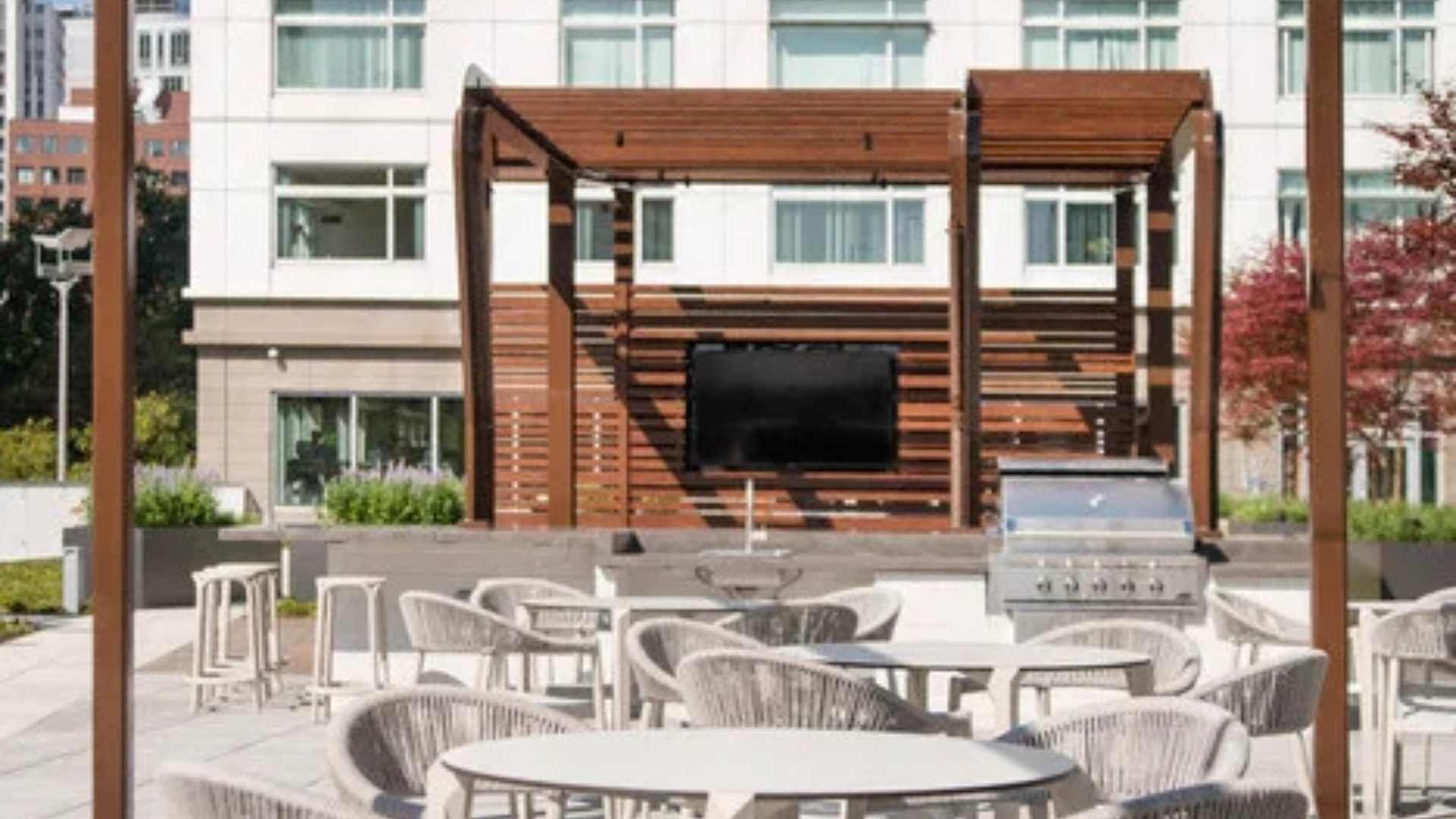 An outdoor seating area with tables and chairs, a wooden pergola structure, and a flat-screen TV mounted on the pergola.