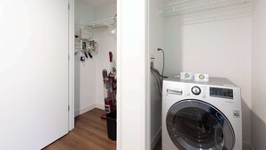 A laundry room with a front-loading washing machine, a vacuum cleaner, and a shelving unit with various items. - Luxury Apt Near Charles River with Sky Terrace Access (Cambridge MA)