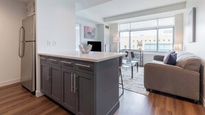 A modern kitchen with grey cabinets, a stainless steel refrigerator, and a central island. There is a grey sofa and a glass table in the living area. - Luxury Apt Near Charles River with Sky Terrace Access (Cambridge MA)