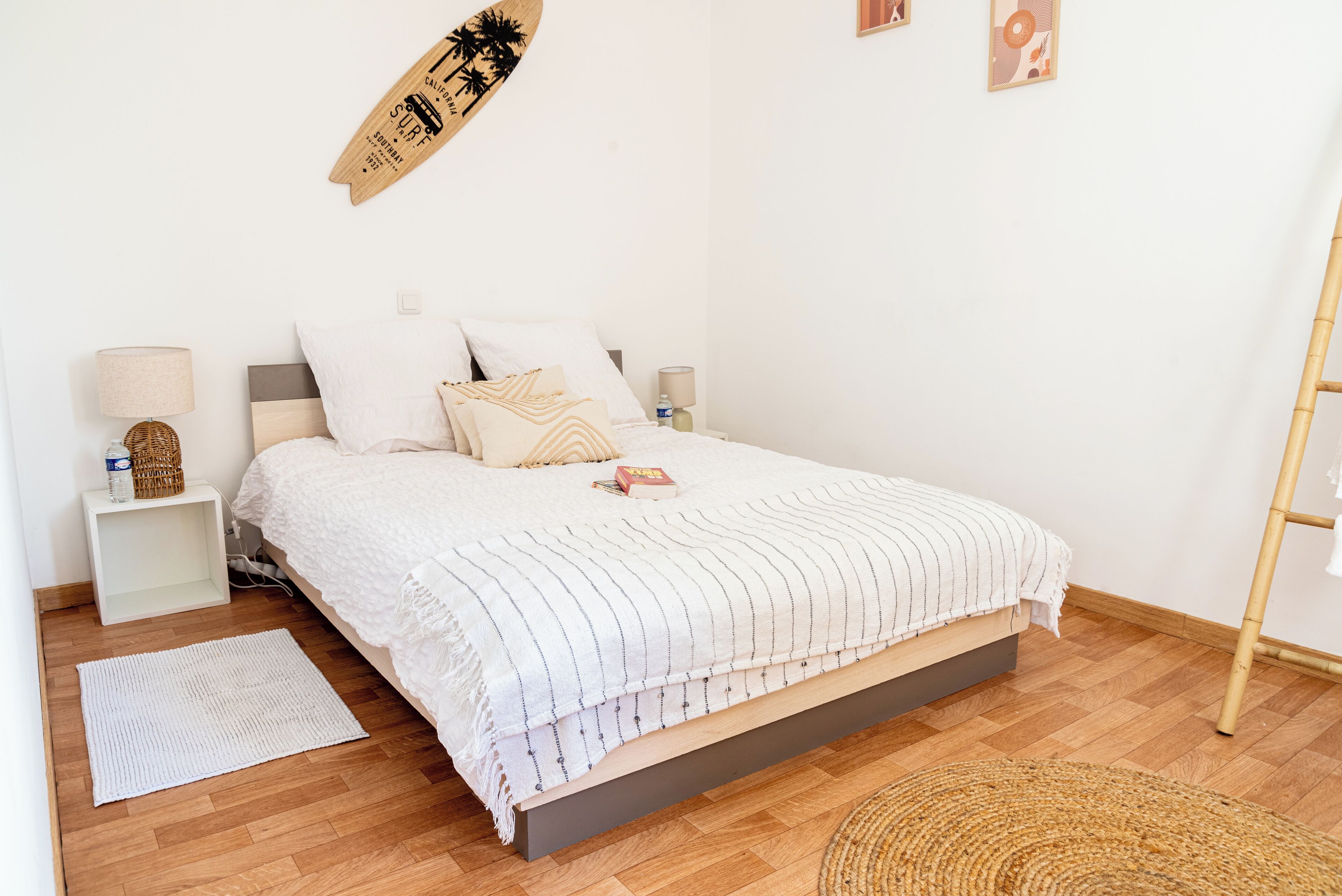 Een netjes opgemaakt slaapkamer met een bed, nachtkastjes, een bord aan de muur en een geweven mat op de vloer.