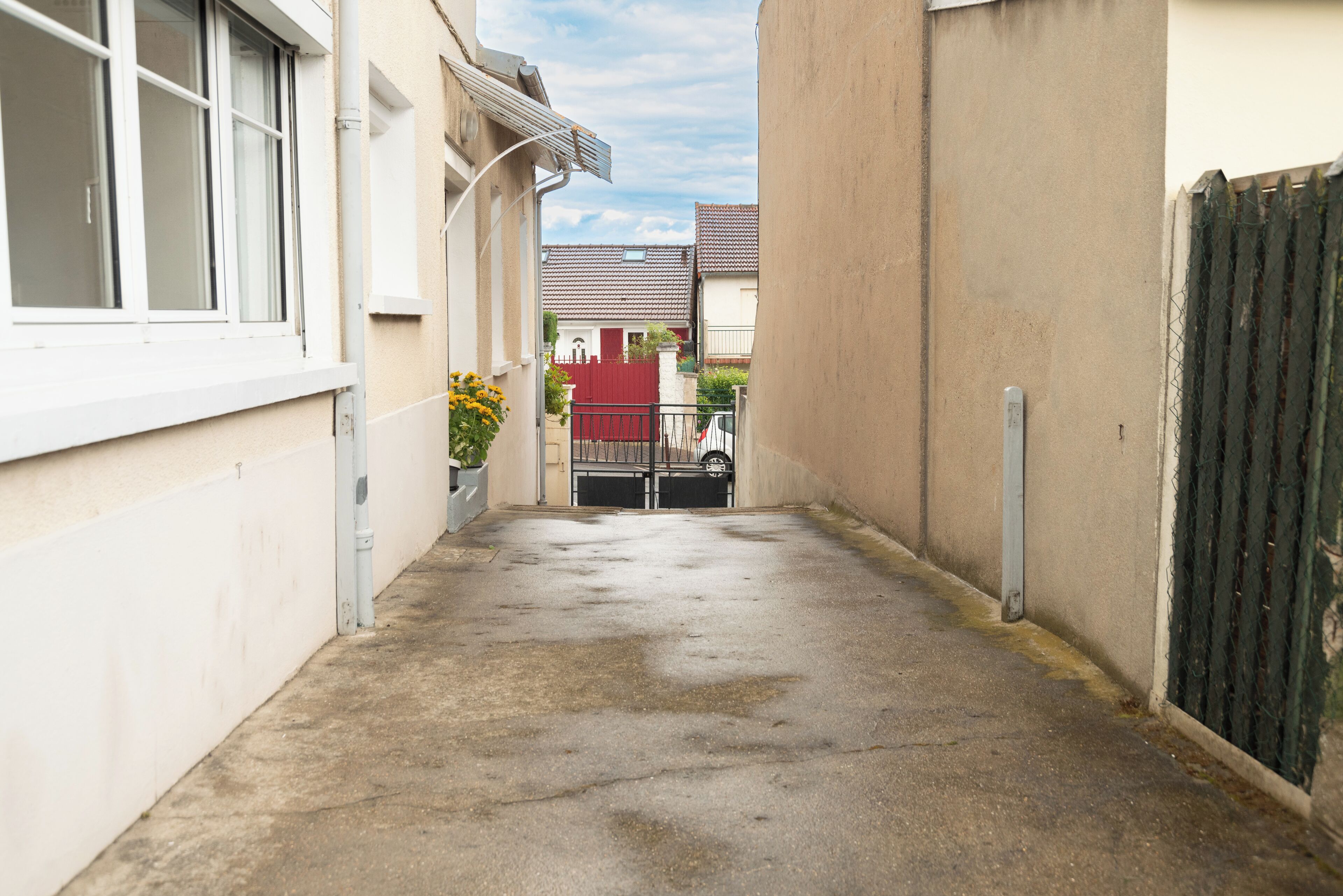 A narrow alleyway with a concrete floor, a red gate, and a white building with a window on the left.