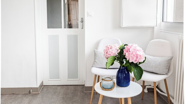 A room with a white door, a white chair, a small round table with a blue vase, and a white furry rug.