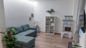 Een moderne woonkamer met een hoekbank, een bijzettafeltje met een lamp en een plant, een witte boekenkast met diverse spullen, en een televisie op een dressoir.
