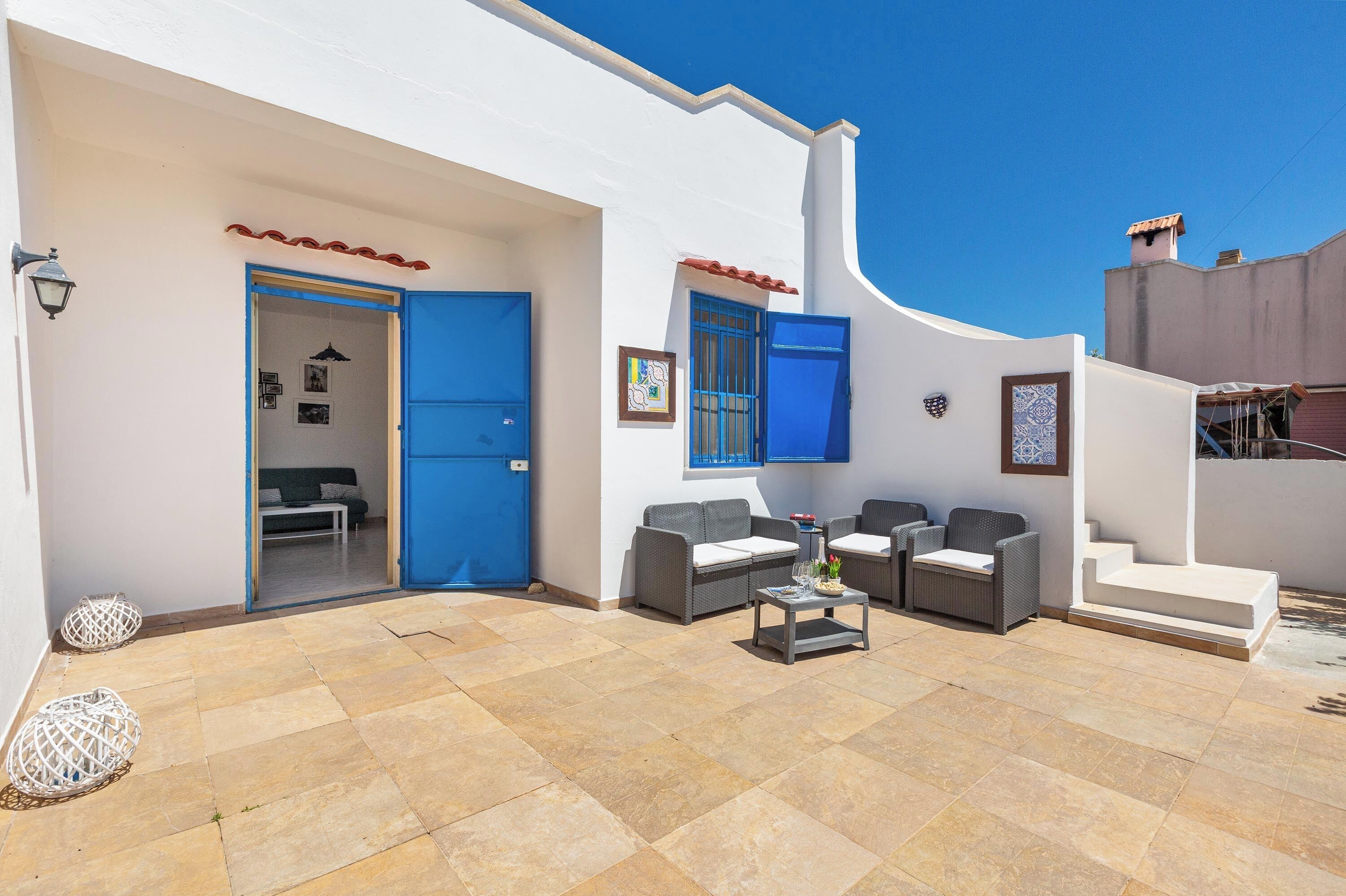 Un edificio blanco con puertas y ventanas azules, un patio con pavimento de baldosas y mobiliario exterior, y una escalera que conduce a un nivel superior.