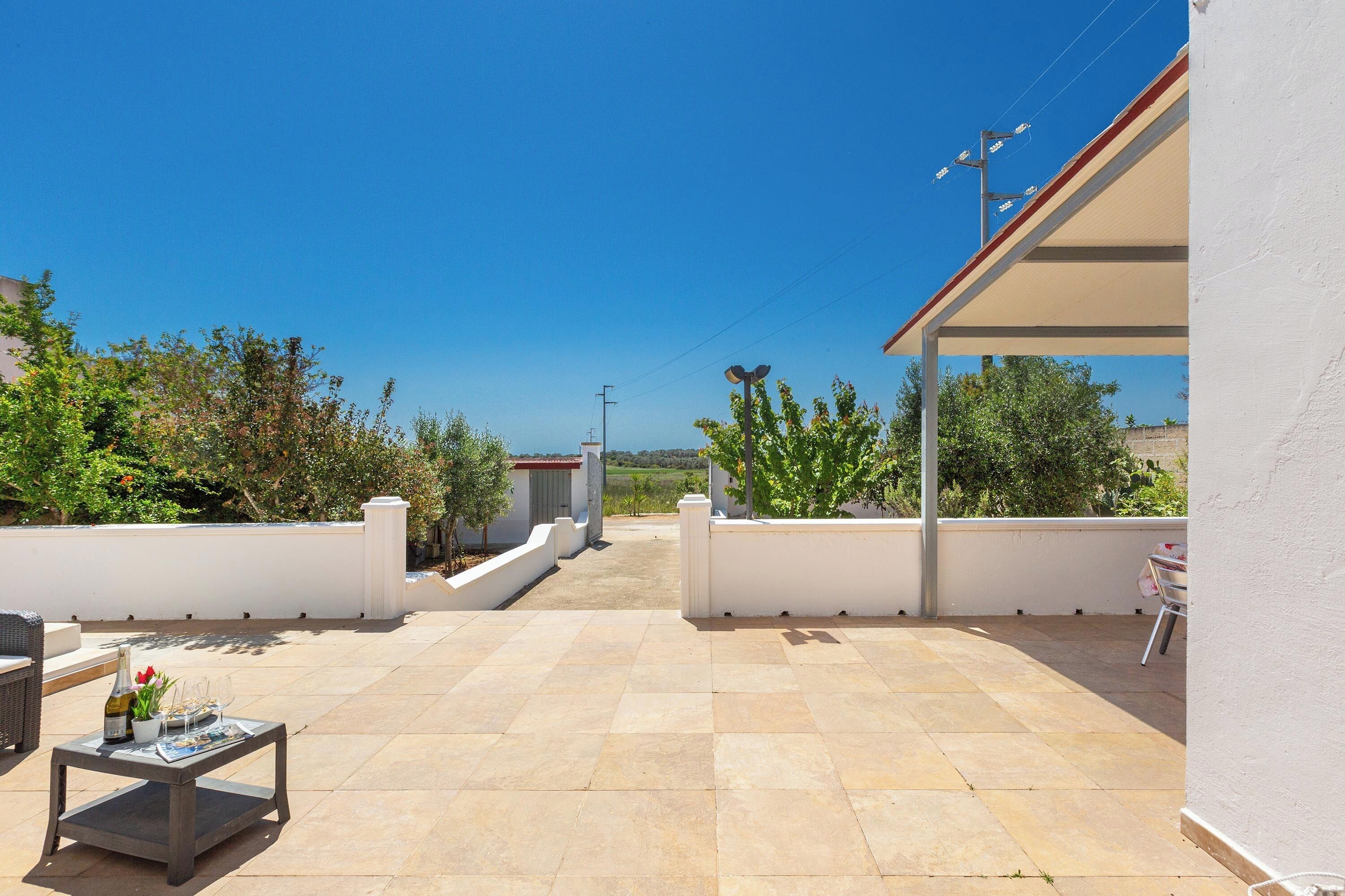 A rooftop terrace with a tiled floor, a table with a bottle and glasses, and a view of the surrounding greenery and buildings.