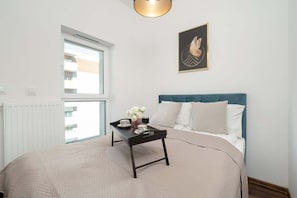 Un dormitorio con cama, mesita de noche con bandeja, ventana y un cuadro colgado en la pared.