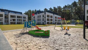 Ein Spielplatz mit Rutsche und Schaukeln, umgeben von einem Gebäude mit vielen Fenstern und blauem Himmel.