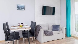 A modern living room with a dining area, a flat-screen TV mounted on a white brick wall, and a sofa with a blanket. - Spacious Apartment | Parking, Balcony and Mountain View (Wisla)