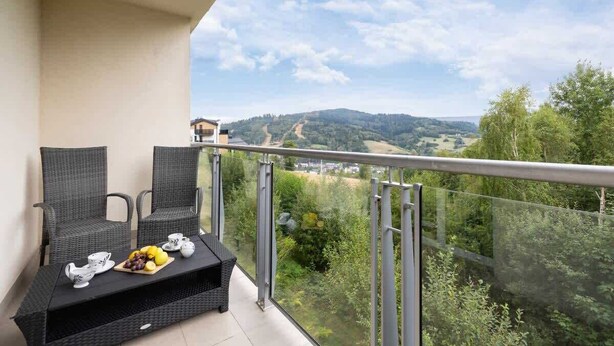 A balcony with wicker furniture, a table with a fruit plate, and a scenic view of hills and trees. - Spacious Apartment | Parking, Balcony and Mountain View (Wisla)