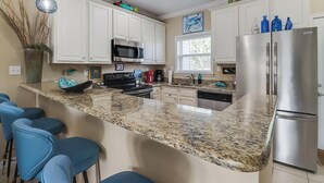 Una cocina con armarios blancos, encimera de granito, refrigerador de acero inoxidable y taburetes de bar azules.