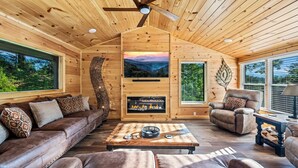 Ein gemütliches Blockhaus-Interieur mit Holzbalkendecke, Kamin, Sofa, Sessel und Wandfernseher.