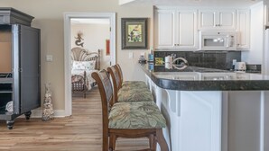 A kitchen with white cabinets, a granite countertop, and a microwave. There is a wooden chair with a cushion in the foreground.