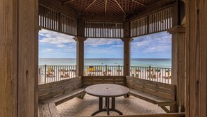 A wooden gazebo with a round table and benches, overlooking a beach with umbrellas and people.
