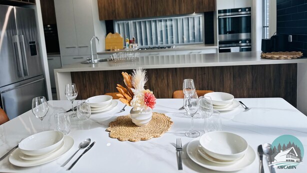 Een moderne keuken met roestvrijstalen apparatuur, een eethoek met een witte tafel gedekt voor vier personen, en een zicht op de keuken door een glazen wand.