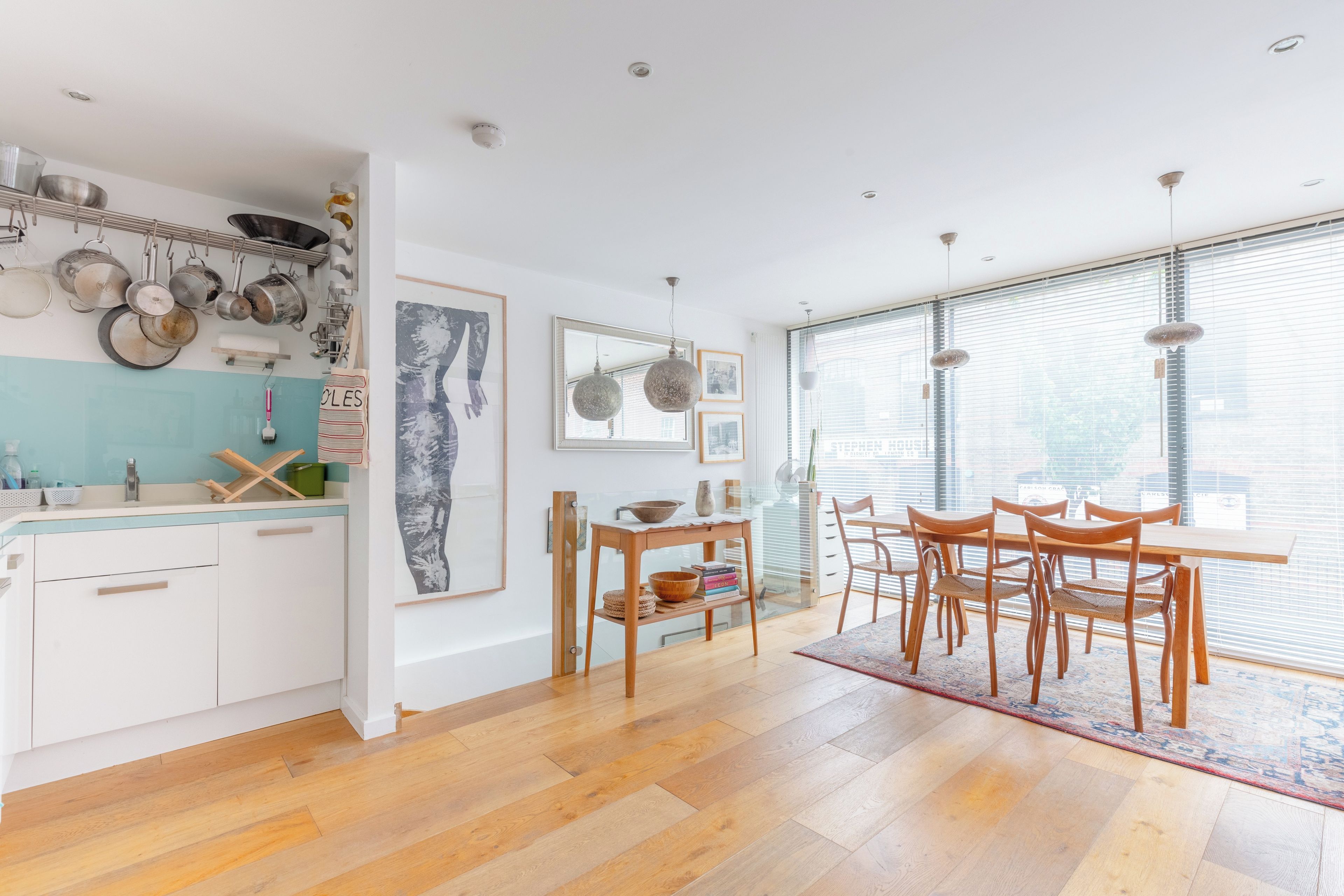 A modern kitchen with wooden cabinets, a dining area with a table and chairs, and a large window with blinds.