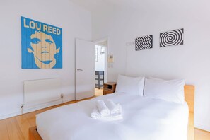 Une chambre d’hôtel avec un lit, du linge de lit blanc, une affiche au mur et une porte donnant sur une autre pièce.