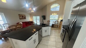 A modern kitchen with white cabinets, a black countertop, and stainless steel appliances. There is a dining area with wooden chairs and a red sofa.