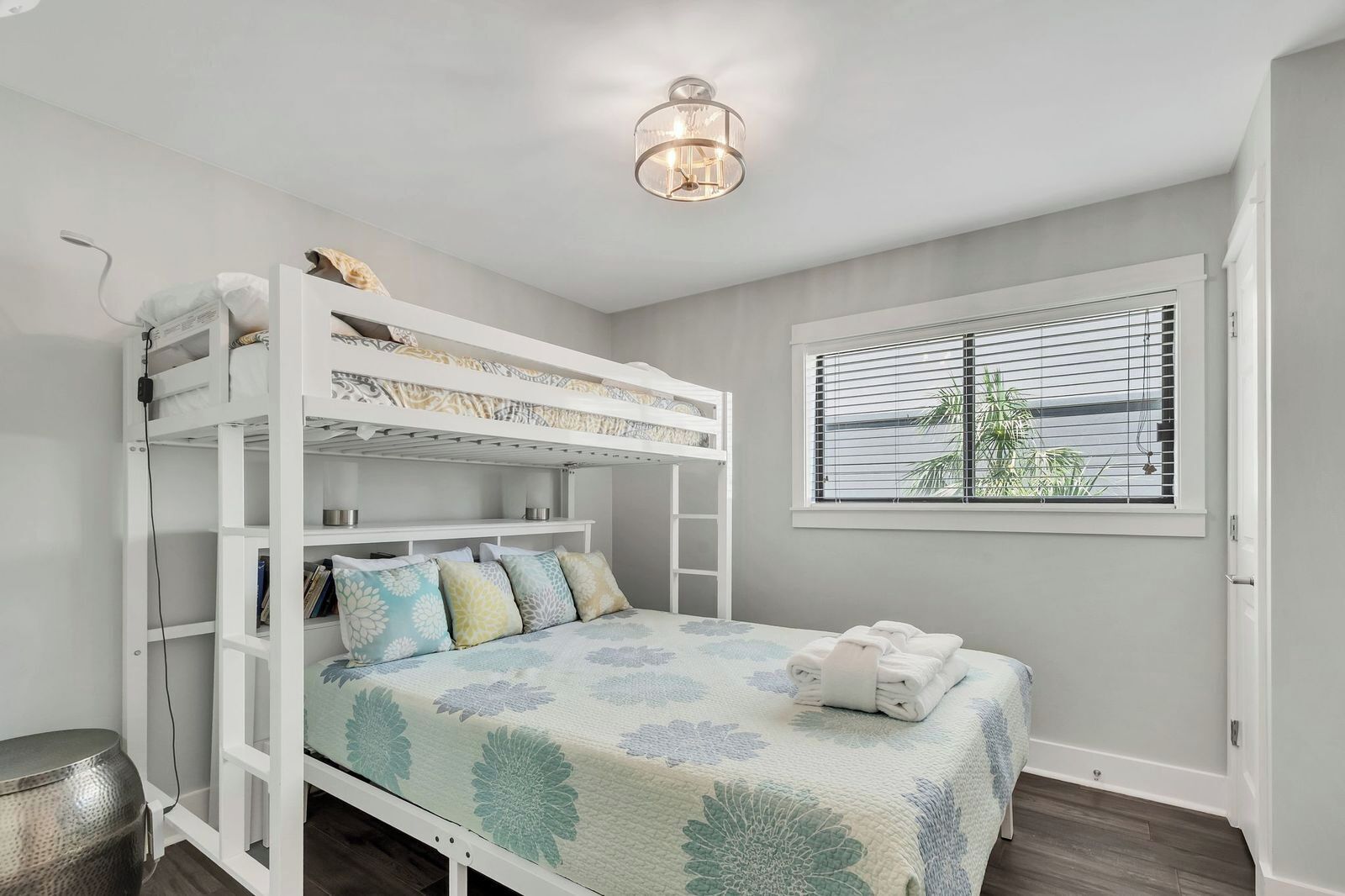 A bunk bed with a floral bedspread, a bedside table, a lamp, and a window with blinds.