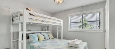 A bunk bed with a floral bedspread, a bedside table, a lamp, and a window with blinds.
