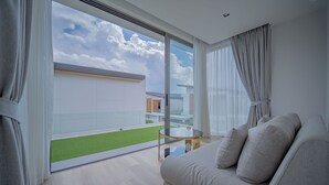 A modern living room with a large sliding glass door leading to a balcony with a green lawn. There is a sofa and a small round table with a glass top. - Zenithy 3BR Villa with Pool by NLA (Choeng Thale)