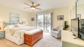 Une chambre à coucher comprenant un lit, une commode en bois, un bureau, une chaise, un ventilateur de plafond et une grande fenêtre.