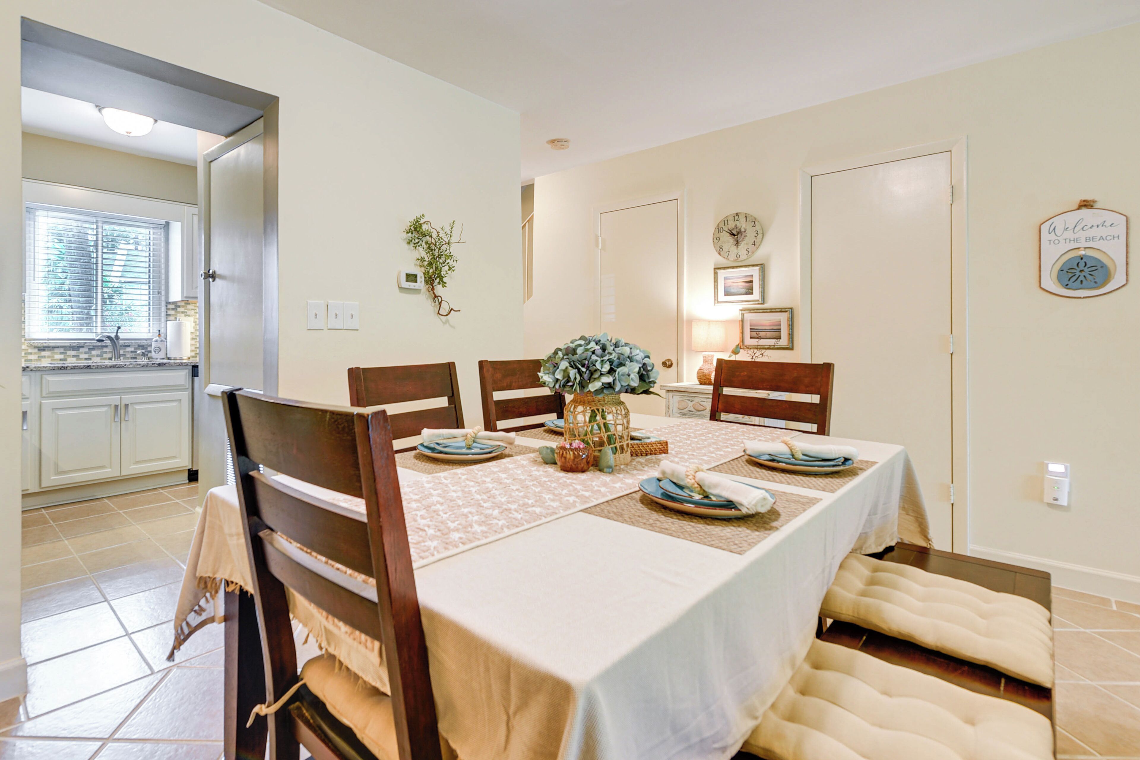 A dining area with a table set for four, surrounded by wooden chairs. A vase with flowers is placed on the table, and there is a clock on the wall.