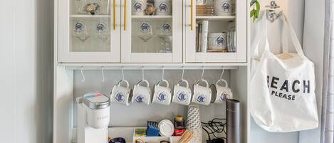A kitchen with a coffee station, a cabinet with decorative mugs, and a hanging tote bag.