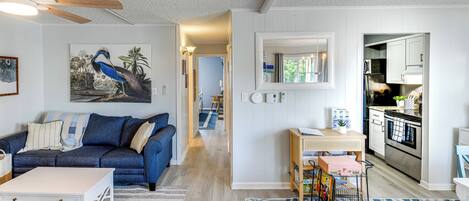 A modern living room with a blue sofa, a white coffee table, and a kitchen area visible through an open doorway.