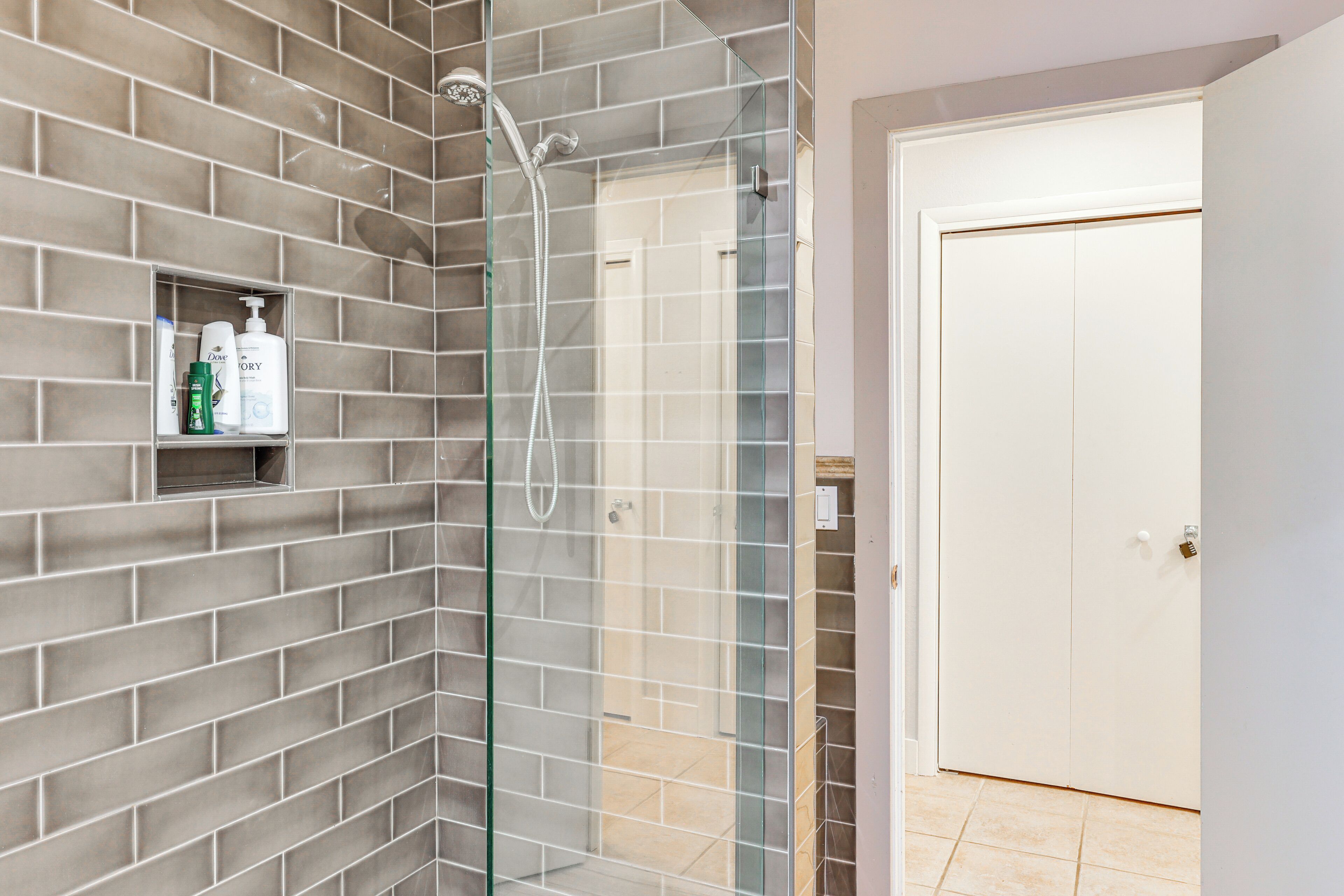 A shower with a glass door, a shelf with toiletries, and a white door in the background.