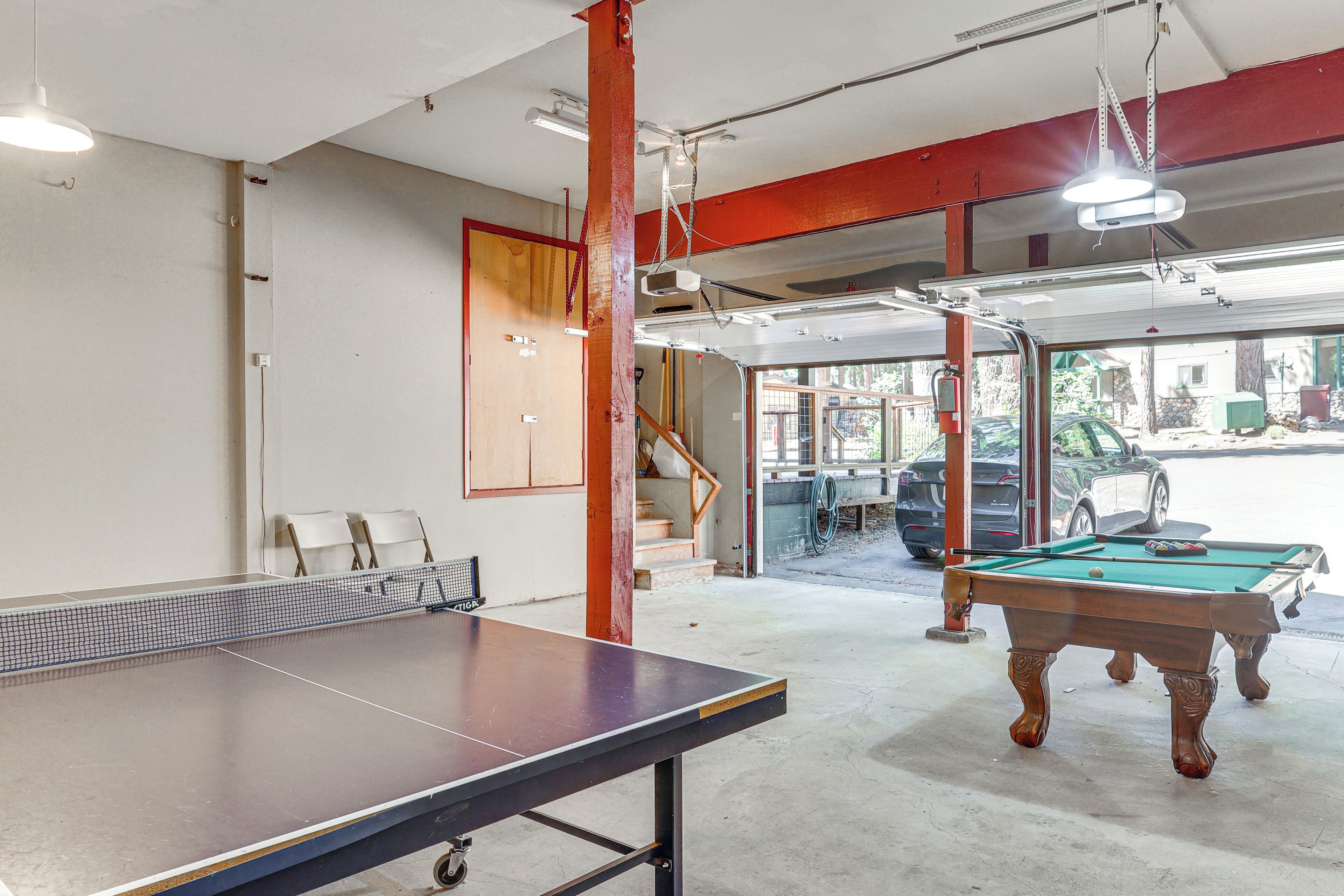 Une table de billard et une table de ping-pong se trouvent dans une pièce dont le sol est en béton, agrémentée d’éléments en bois et comportant un escalier menant à un niveau supérieur.