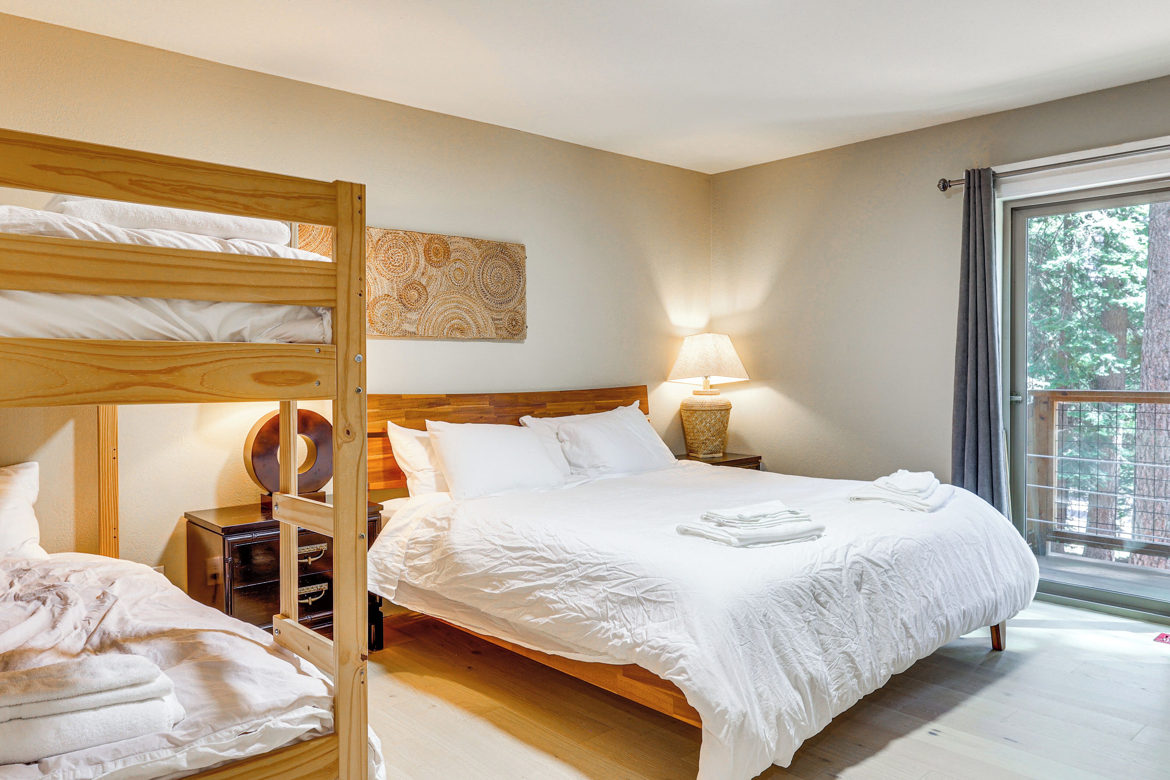 A bedroom with a bunk bed, a single bed, a bedside lamp, and a window with a view of trees.