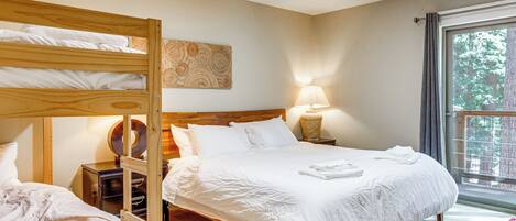 A bedroom with a bunk bed, a single bed, a bedside lamp, and a window with a view of trees.
