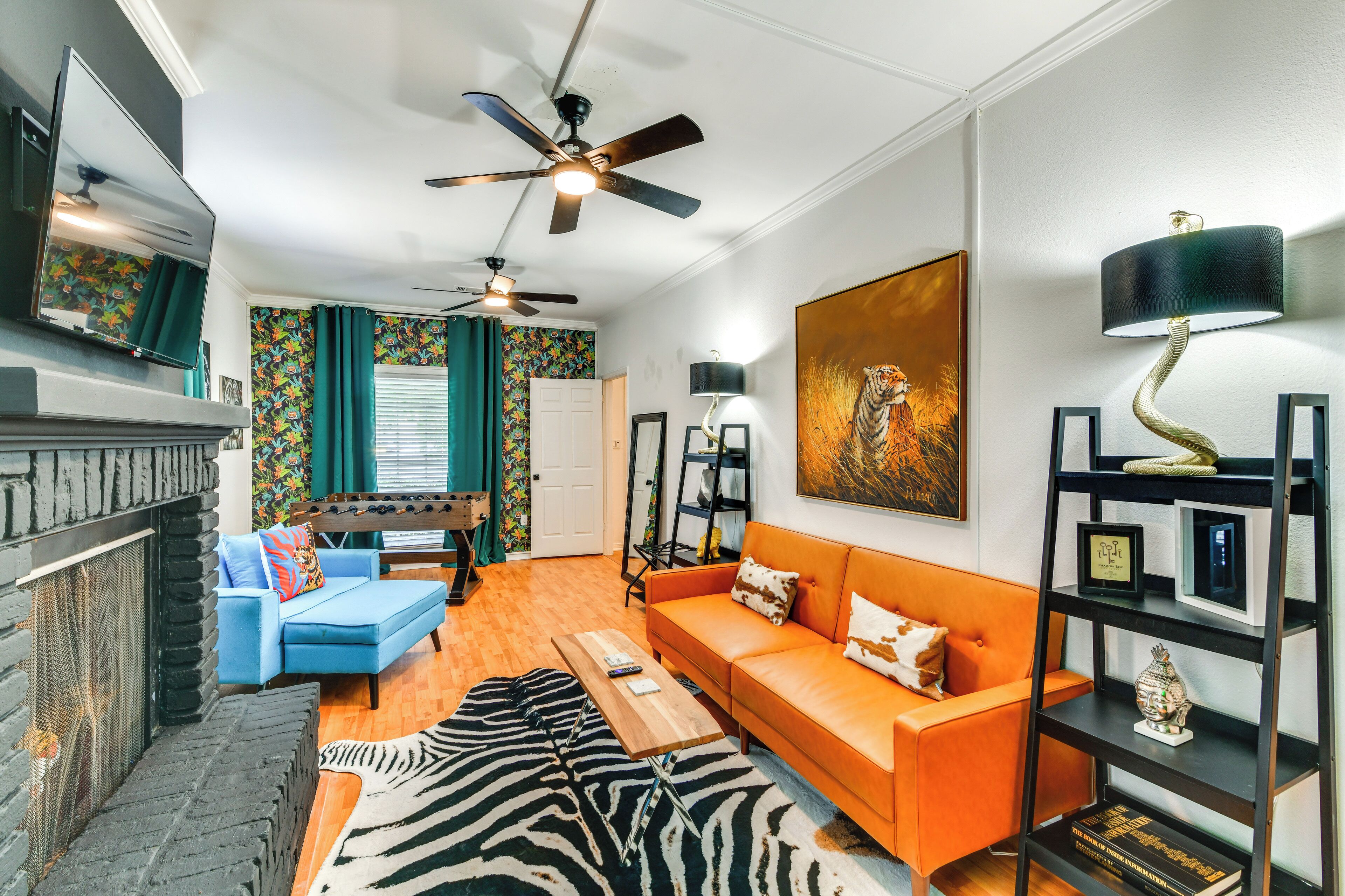 A modern living room with a fireplace, blue sofa, orange sofa, zebra rug, and a ceiling fan.