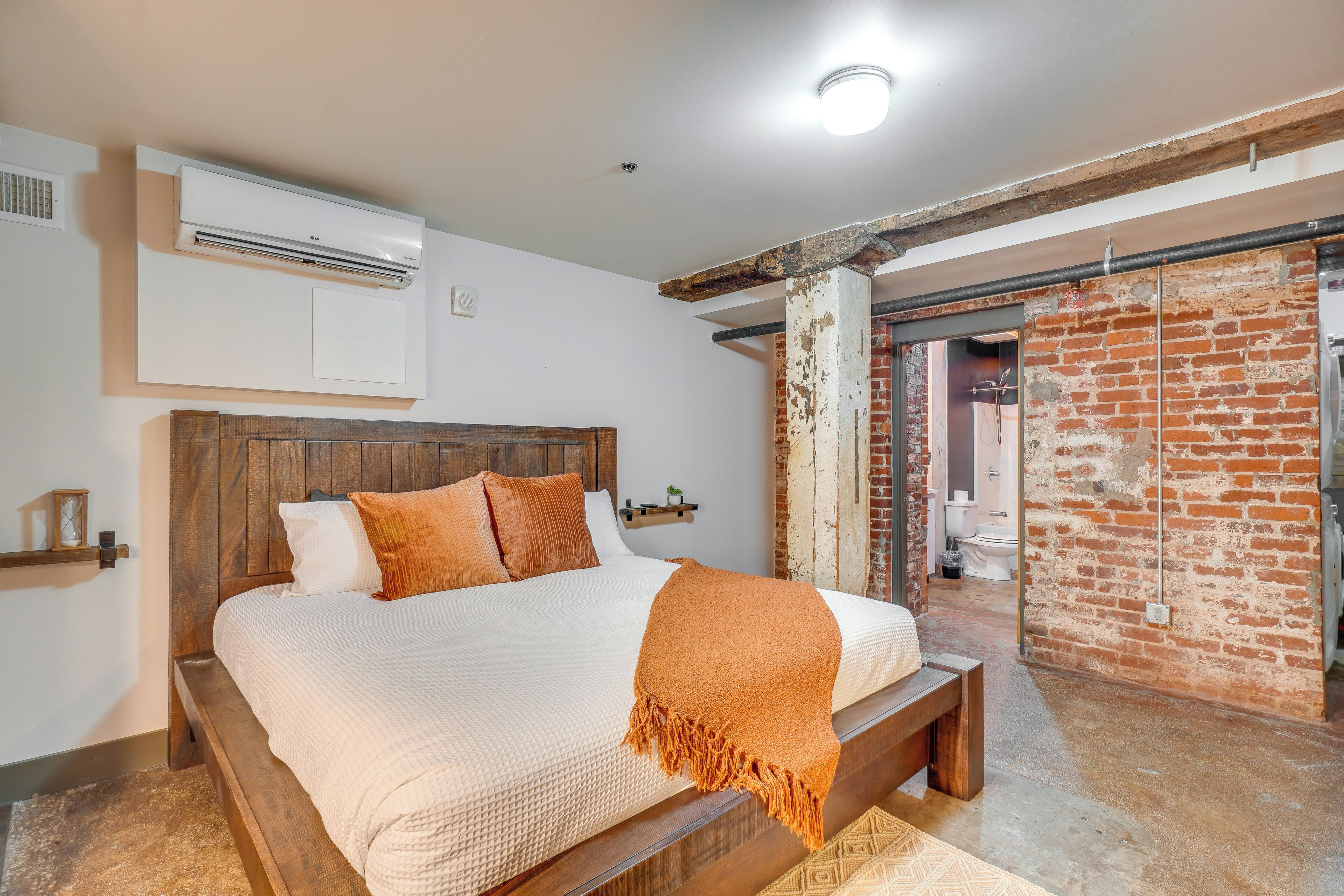 A bedroom with a wooden bed, a white wall-mounted air conditioner, and a brick wall.