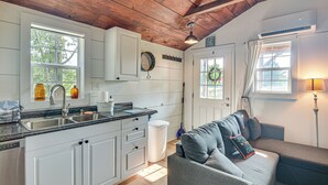 A kitchen with white cabinets, a sink, and a stove. There is a gray couch with pillows and a lamp in the living area.