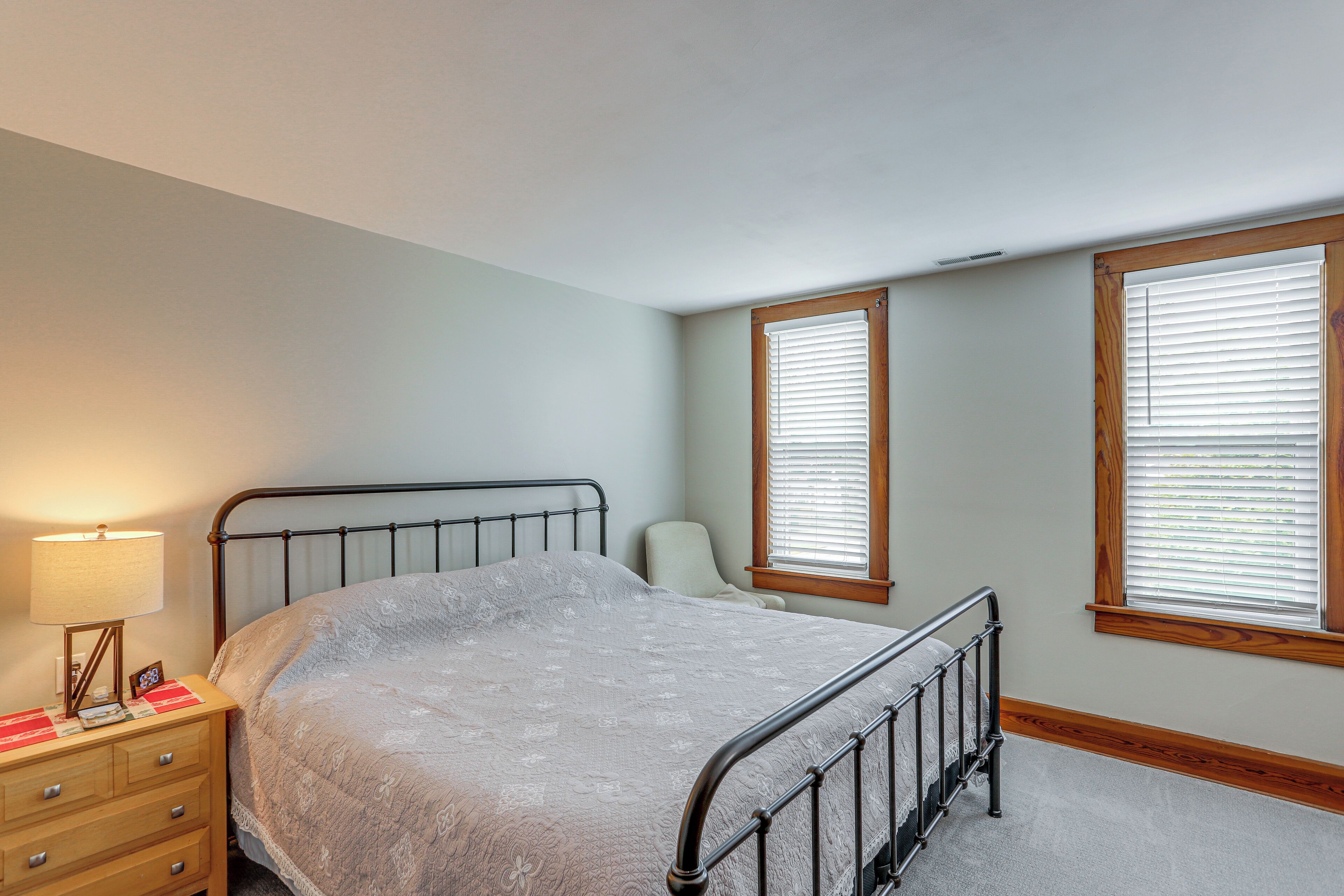A bedroom with a bed, a nightstand, a lamp, and two windows with blinds.