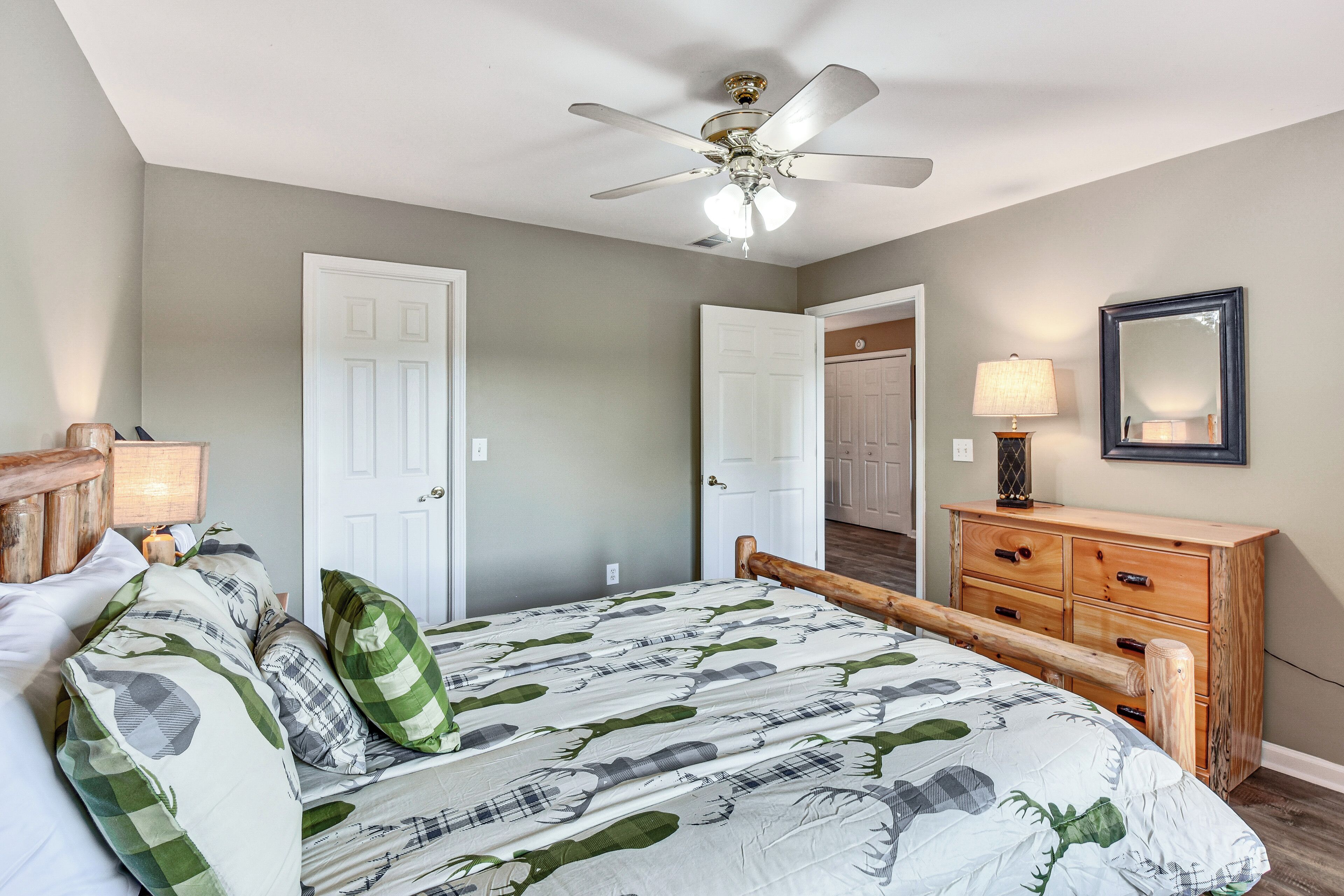 A bedroom with a bed, a wooden dresser, a ceiling fan, and a door leading to another room.