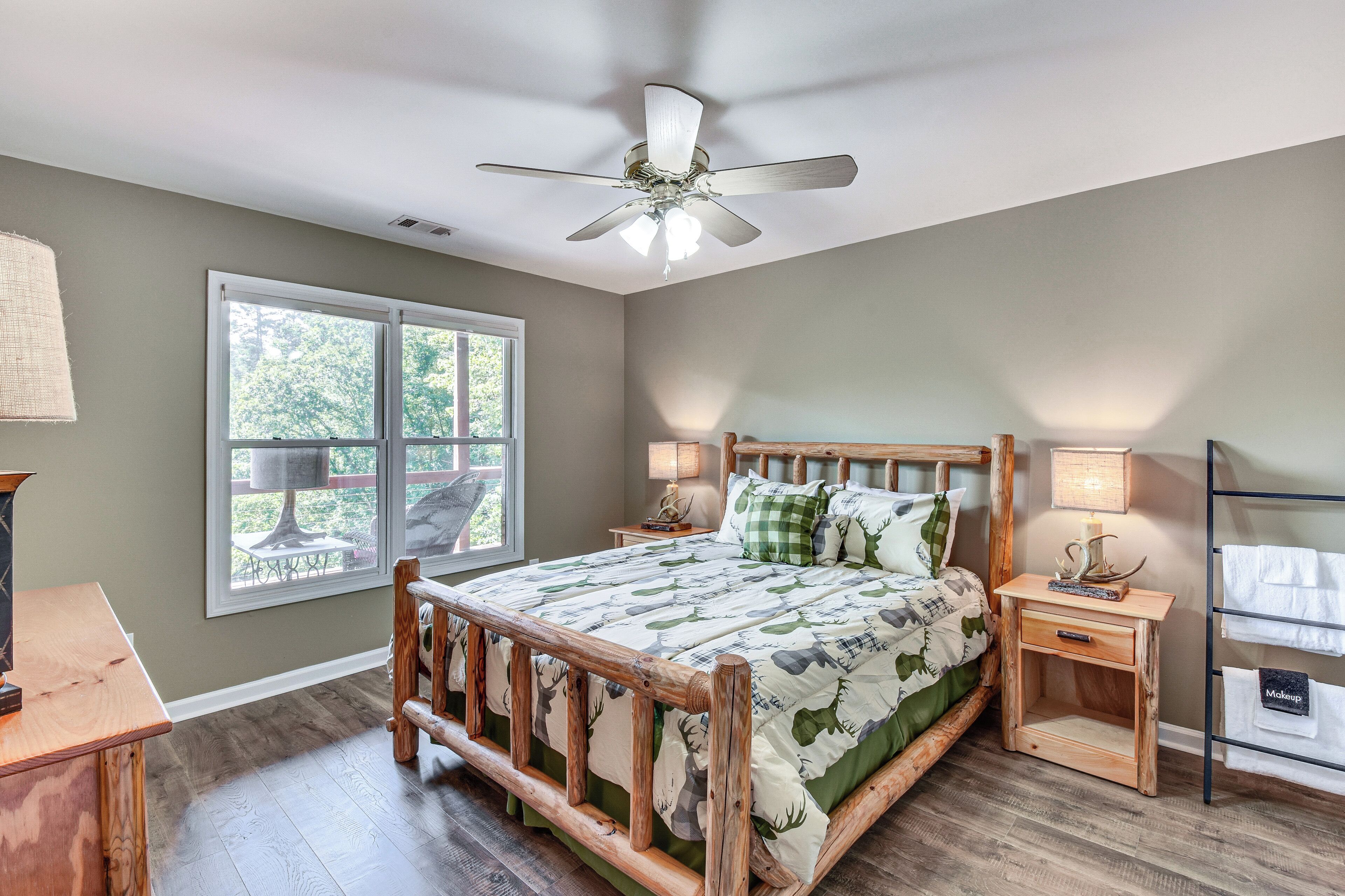 A bedroom with a wooden bed frame, a ceiling fan, a window with a view of greenery, and a bedside table with a lamp.