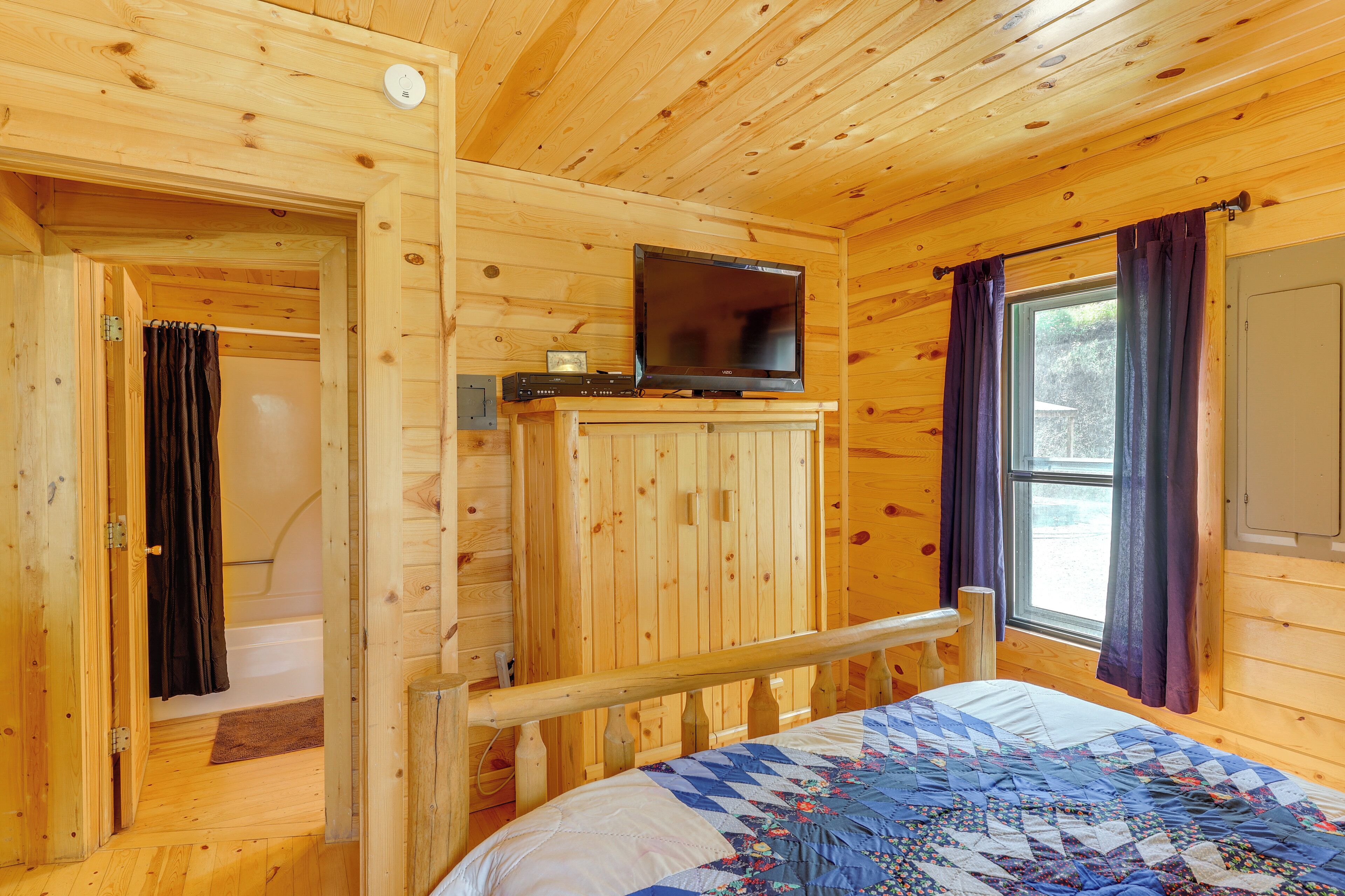 Habitación tipo cabaña de madera con una cama, un televisor sobre un mueble, una ventana con cortinas y una puerta que da a un baño.