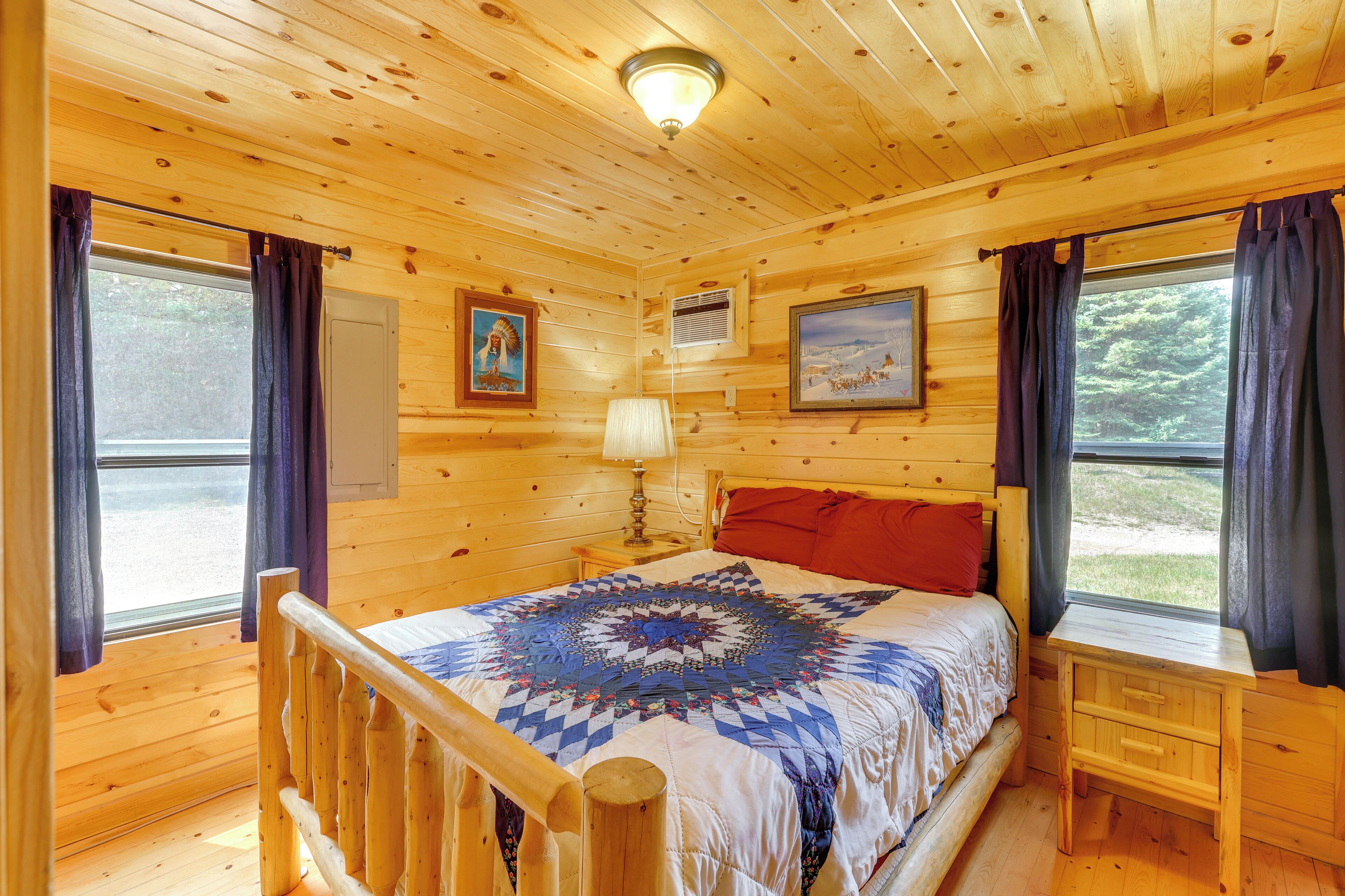 Un dormitorio de cabaña de madera con una cama, una mesita de noche, una ventana con cortinas y una lámpara en el techo.