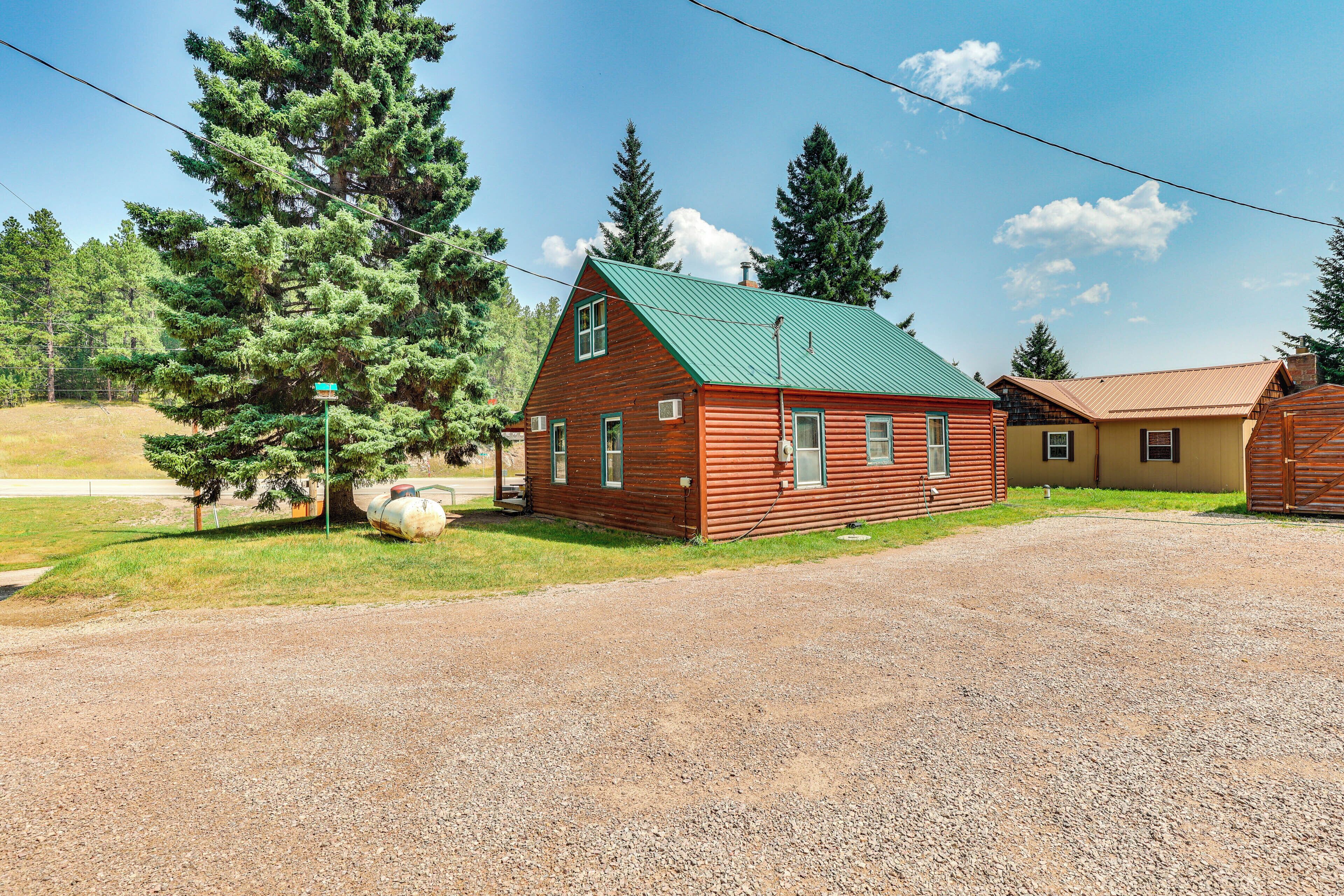 A rustic log cabin with a green roof, a gravel driveway, and a small adjacent building.