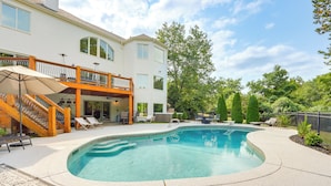 Ein großer, gebogener Swimmingpool mit Holzterrasse und Liegestühlen, umgeben von einem mehrstöckigen Haus mit einem Balkon.
