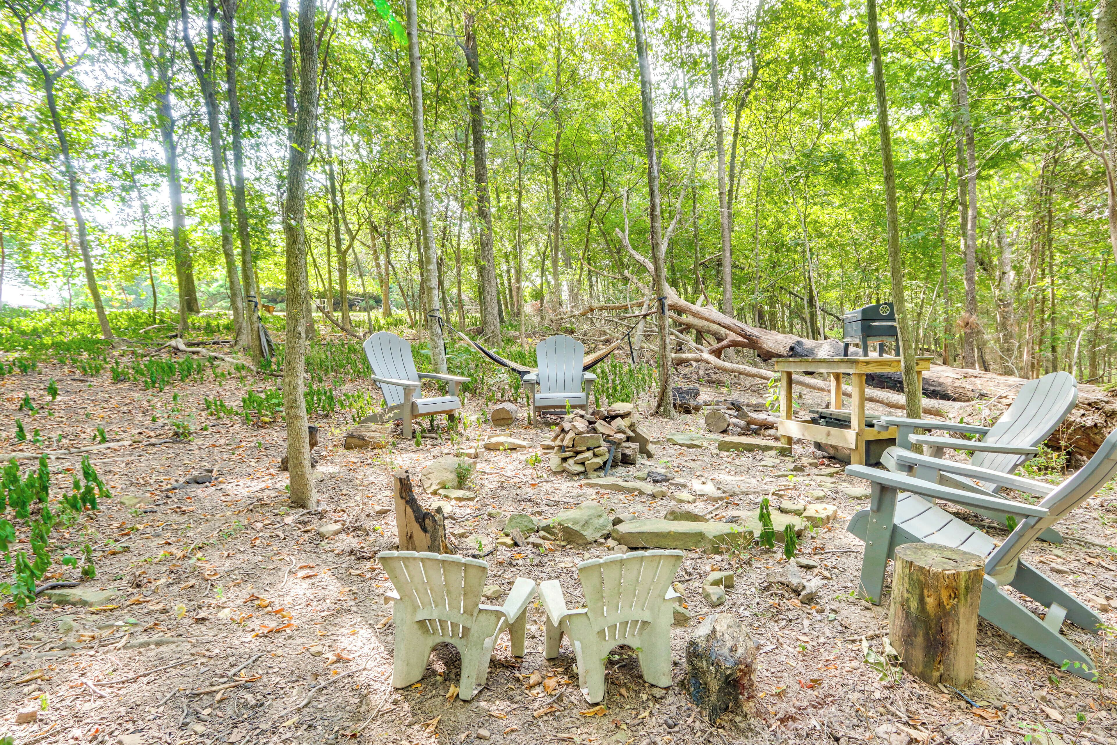 A forested area with outdoor seating including Adirondack chairs and a table, surrounded by trees and natural ground cover.