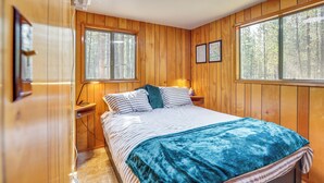 Un dormitorio con paneles de madera, una cama, dos ventanas y una mesita de noche con lámpara.