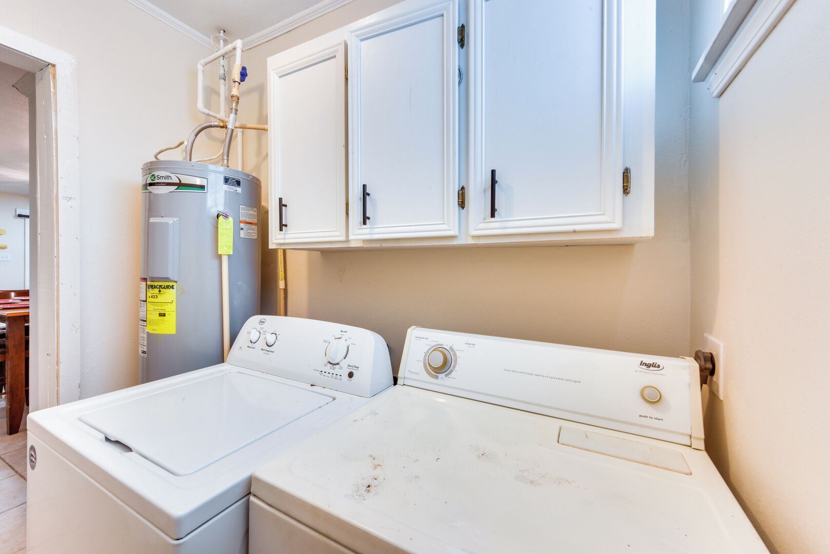 A laundry room with two white washing machines, a water heater, and white cabinets above.