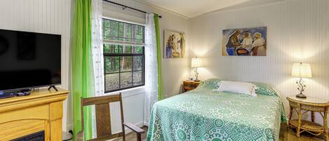 Un dormitorio con cama, televisión, silla, mesita de noche, lámpara y un cuadro en la pared.