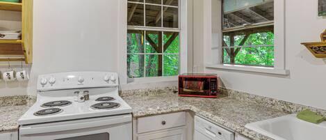 A kitchen with a white stove, a microwave, and a sink. There are windows with views of trees.
