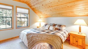 A bedroom with a sloped ceiling, a bed with a checkered bedspread, two bedside lamps, and a wooden nightstand. - Deck w/ Mountain Views: Cabin in Bear Valley! (Arnold)